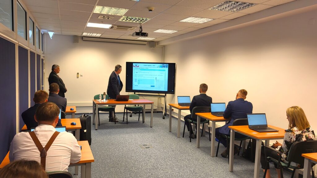 Fot. 2. Zdjęcie przedstawia salę egzaminacyjną z członkami Komisji egzaminacyjnej i osobami przystępującymi do egzaminu siedzącymi pojedynczo przed laptopami.