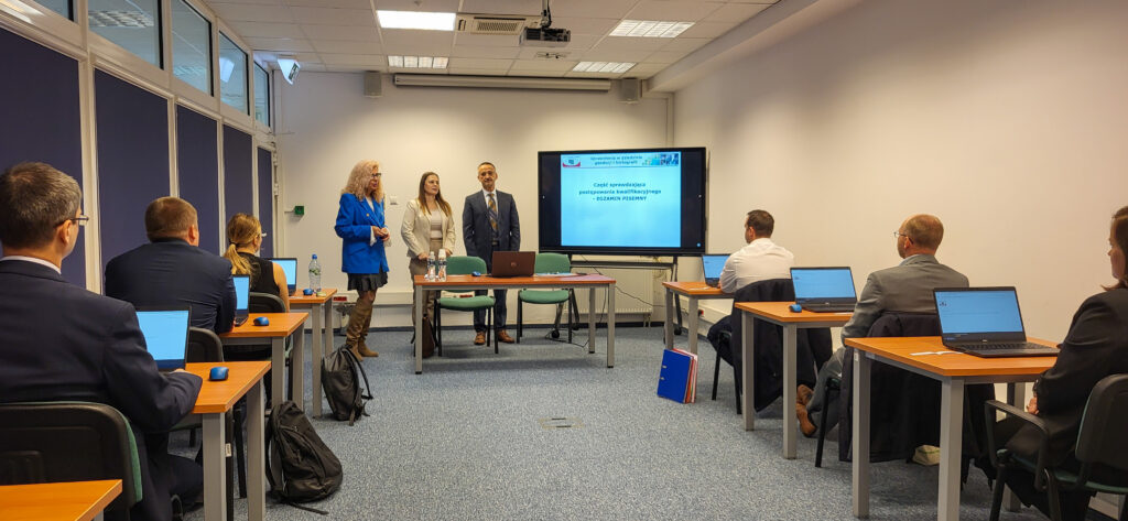 Fot. 3. Zdjęcie przedstawia salę egzaminacyjną z członkami Komisji egzaminacyjnej i osobami przystępującymi do egzaminu siedzącymi pojedynczo przed laptopami.